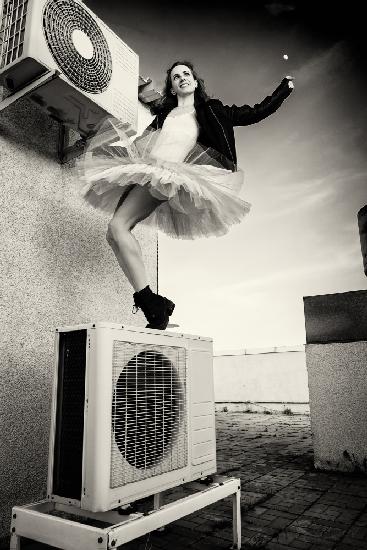 a ballerina in tutu, jacket and boots climbed on the air conditioner and poses against the sky